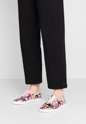 Personne portant un pantalon noir et des baskets à lacets à motifs floraux avec des semelles blanches, debout sur un sol gris lisse.