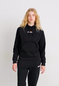 Manta preta com um bolso frontal, capuz com cordão, e logótipo de marca em branco e vermelho. Combinada com calças de fato pretas com logótipo.