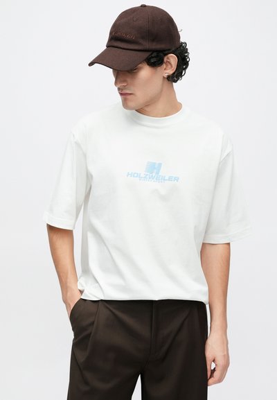 Tricou alb cu mâneci scurte și grafică albastră „HOLZWEILER WINTERSPORT”, asortat cu pantaloni maro și șapcă maro. Croială simplă, lejeră.