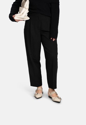 Persona che indossa pantaloni neri e un maglione nero testurizzato, che tiene in mano una borsa beige piegata, indossa ballerine beige appuntite con fibbie e borchie.