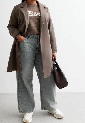 Persoon draagt een taupekleurige jas over een bruine trui, lichte jeans met wijde pijpen, witte sneakers, houdt een donkerbruine handtas vast, staat tegen een witte achtergrond.