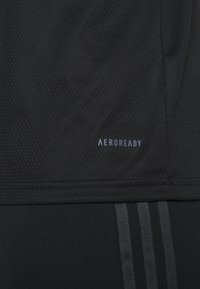 Primo piano di un tessuto nero testurizzato con il logo "AEROREADY" e vista parziale di pantaloni neri con tre strisce verticali.
