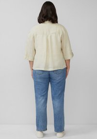 Lichtbeige linnen shirt met opgerolde mouwen, gecombineerd met mid-blauwe rechte jeans, weergegeven van de achterkant tegen een effen achtergrond.