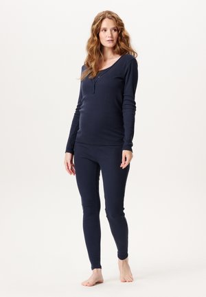 Kvinde med langt, bølget hår iført marineblå langærmet top og matchende leggings, stående barfodet på en ensfarvet lys baggrund.