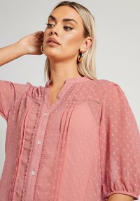 Blusa rosa chiaro con pois testurizzati, chiusura a bottoni sul davanti, colletto arricciato e maniche a palloncino corte con dettagli in pizzo.