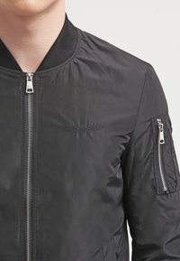 Svart bomberjacka med en slät yta, framdragkedja och ribbad krage. Har sidofickor och en broderad logotyp på bröstet.