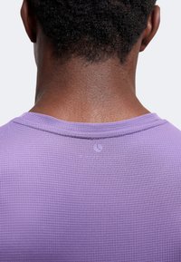 Πλάτη ενός ατόμου με κοντά σγουρά μαλλιά που φοράει μια υφασματική μωβ μπλούζα με ένα μικρό λογότυπο ημισελήνου στο λαιμό.