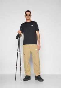 Tricou negru cu logo, pantaloni scurți bej, și încălțăminte de drumeție negre. Ținând bețe de trekking, poartă ochelari de soare bej. Design simplu și funcțional.