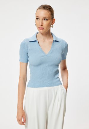 Vrouw draagt een lichtblauw kort mouw geribd gebreid topje met kraag en witte hoge taille broek, staand met één hand in de zak.