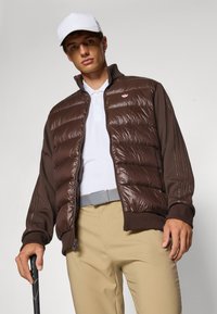 Smeđa pufasta jakna s rebrastim rubom i ovratnikom, uparena s bijelom polo majicom i bež hlačama, uz sivi remen i golf štap.