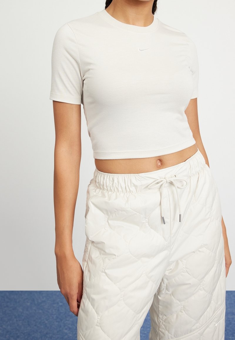 Jasnobeżowy krótki t-shirt o okrągłym dekolcie i krótkich rękawach, z małym białym logo Nike. W zestawie z białymi pikowanymi spodniami.