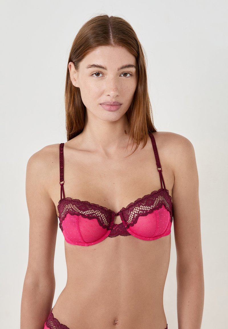Giovane donna con capelli lisci castano chiaro che indossa un reggiseno in pizzo rosa e bordeaux, in piedi contro uno sfondo chiaro, con lo sguardo rivolto in avanti.
