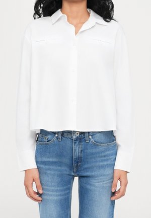 Nainen pukeutuneena valkoiseen pitkähihaiseen kauluspaitaan, joka on puettu keskikorkeavyötäröisten, sinisten farkkujen yli. Hän seisoo yksinkertaista taustaa vasten.