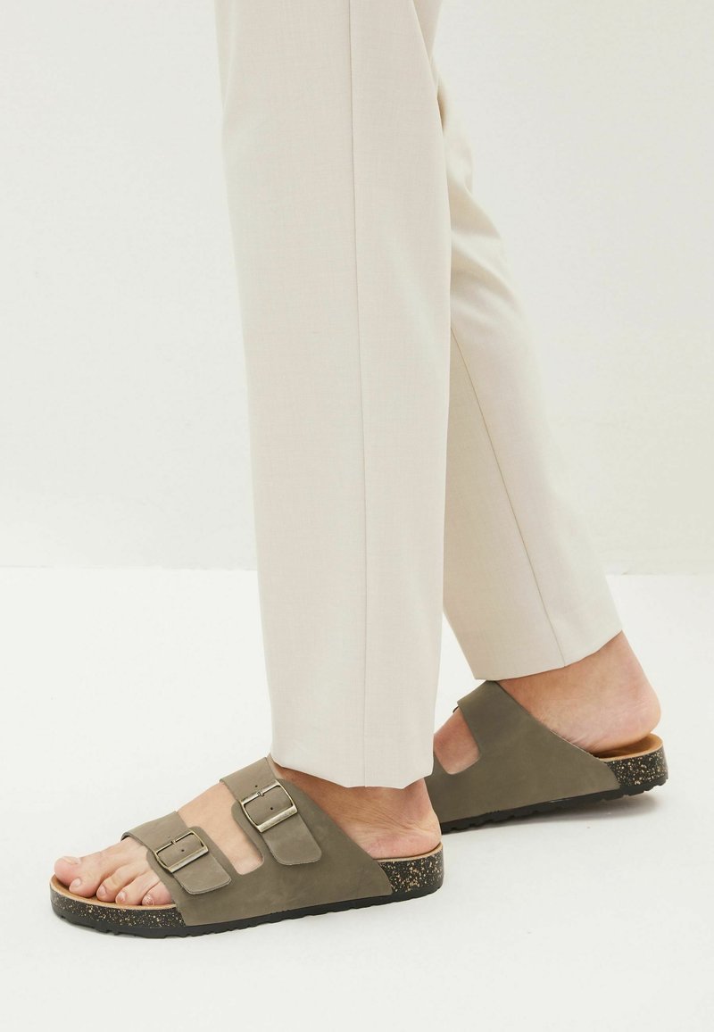 Zapatos planos de ante en color verde oliva, con dos correas ajustables con hebillas plateadas, una suela de corcho texturizada y un diseño minimalista.
