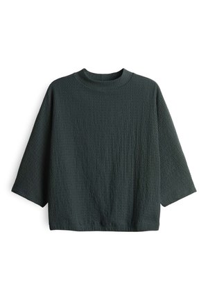 Maglione lungo a maniche lunghe di un verde scuro con collo rotondo e vestibilità ampia, disteso su uno sfondo bianco.