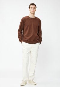 Brązowy długi rękaw sweatshirt z prążkowanym, okrągłym dekoltem, zestawiony z białymi spodniami cargo z bocznymi kieszeniami, noszony z jasnymi sneakersami.