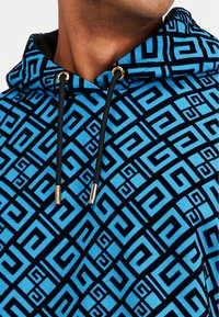 Modrá a černá mikina s kapucí s geometrickým vzorem, černými stahovacími šňůrkami a zlatými kovovými doplňky. Měkká textura a klasický střih.