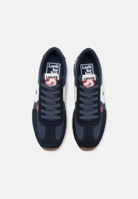 Ett par marinblå och vita sneakers från Levi's med snörning, märkesetiketter på tungan och sidan, sedd uppifrån mot en vit bakgrund.