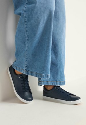 Personne portant un jean bleu à jambes larges et des baskets basses bleu marine à semelles blanches, debout devant un fond clair.