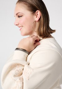 Crèmekleurige trui met versierde pailletten op de mouw. Model draagt zilveren creolen en gelaagde armbanden, wat textuur en detail benadrukt.