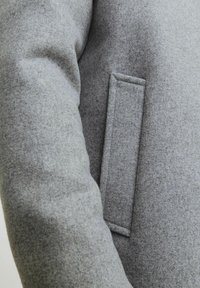 Abrigo de mezcla de lana gris con una superficie texturizada. Presenta un discreto bolsillo de parche en la manga y una silueta ajustada.