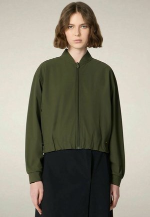Donna con i capelli corti e castani che indossa una giacca bomber verde oliva con zip e una gonna nera su uno sfondo neutro.