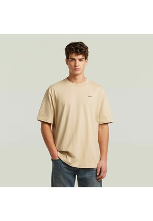Giovane uomo con capelli ricci, che indossa una t-shirt beige oversize e jeans blu, in piedi con una mano in tasca davanti a uno sfondo neutro.