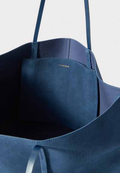 Blue suede tote bag with a smooth finish, featuring a flat interior pocket and thin shoulder straps. Gold embossed brand logo inside.