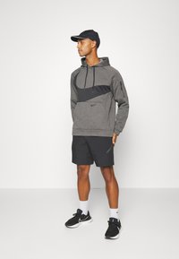 Sudadera gris con acentos negros y bolsillo canguro, combinada con pantalones cortos negros. El modelo lleva zapatillas negras y una gorra negra, de pie sobre un fondo liso.
