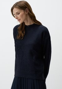 Pull bleu marine foncé en tissu doux, avec un motif floral en relief sur le devant et un col rond classique.