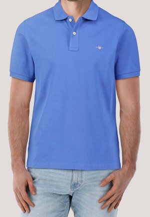 Mężczyzna w niebieskiej koszulce polo z krótkim rękawem i dwoma guzikami oraz jasnoniebieskich dżinsach, stojący z ramionami luźno opuszczonymi wzdłuż ciała.