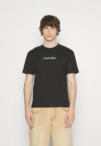 T-shirt en coton noir à manches courtes et col rond, arborant un logo blanc "Calvin Klein" sur le devant.