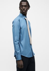 Άνδρας που φοράει μπλε πουκάμισο με μπεζ γραβάτα και μαύρο παντελόνι, κοιτάζει προς τα πλάγια σε απλό λευκό φόντο.