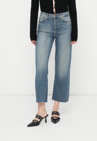 Jeans de ganga azul-claro de perna larga com bainha desfiada; combinados com mules pretos de bico fino com detalhes em metal e tiras.