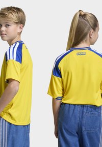 Gul sporttröja med blå detaljer och vita ränder på axlarna, med texten "SVERIGE" tryckt på ryggen. Kortärmad, avslappnad passform.