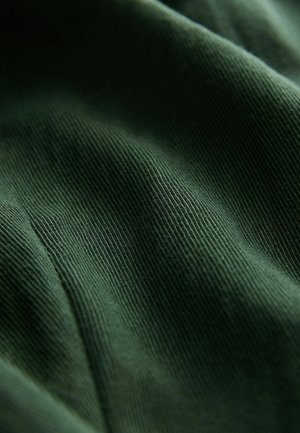 Tumši zaļš ribots audums ar smalku tekstūru un stiepes īpašību. Materiāls parāda smalkas līnijas, kas horizontāli stiepjas pāri virsmai.