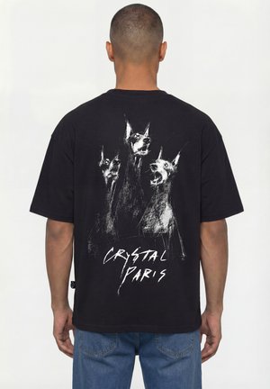 Mężczyzna noszący czarną koszulkę z białym szkicem trzech głów psa rasy Doberman i napisem „Crystal Paris” na plecach.