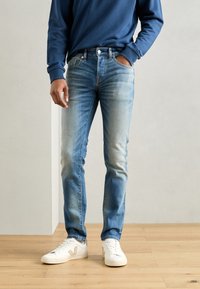 Helle blaue Jeans im Slim-Fit, mit dezenten Verwaschungen, Fünf-Taschen-Design und Messingbeschlägen, kombiniert mit weißen Sneakers.