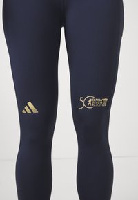 Mornarske legice iz elastičnega materiala, z zlatim Adidasovim logotipom in zlatim besedilom "50 BMW Berlin Marathon" ter grafičnim ikonastim simbolom.