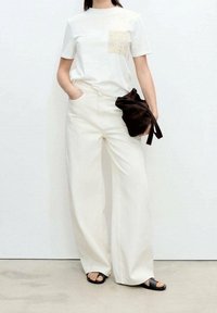 Personne portant une chemise blanche à manches courtes avec une poche en dentelle, un pantalon large blanc, des sandales noires, tenant un sac en tissu noir.