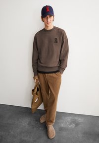 Sweatshirt marron avec un petit logo, pantalon en velours côtelé beige, chaussons gris, et une casquette navy avec un accent en lettre rouge. Tient un sac fourre-tout beige.
