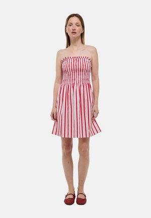 Femme debout portant une robe sans bretelles à rayures verticales rouges et blanches et des chaussures plates rouges, sur un fond clair uni.