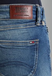 Modré džíny z denimu s hnědým koženým štítkem označeným "TOMMY JEANS" na pase; mírné vyblednutí a malá trojbarevná značka na kapse.