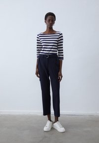 Randig marinblå och vit långärmad topp som matchas med marinblå byxor med bälte och fickor; vita sneakers avslutar outfiten.