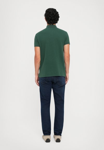 Camisa polo verde con cuello clásico, mangas cortas y dobladillo recto, combinada con jeans de mezclilla azules y calzado de color claro.