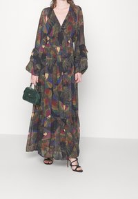 Robe longue en tissu noir transparent avec un motif coloré de plumes. Dispose de manches longues, d'un col en V profond et de détails à volants. Portée avec un sac vert.