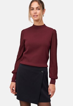Femme portant un pull bourgogne avec des manches bouffantes transparentes à nervures et une mini jupe noire croisée à l'avant avec un bouton sur le côté.