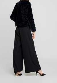 Femme portant un pantalon large noir, des talons hauts noirs, et une veste en fausse fourrure noire texturée, debout devant un fond gris.