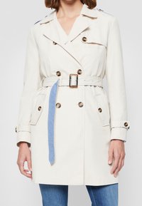 Béžový trenčkot s dvojitým zapínáním, zlatými knoflíky a denimovým páskem. Obsahuje klopový límec a boční kapsy.