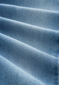 Tejido de mezclilla en azul claro, con una textura sutil que presenta patrones de tejido y pliegues visibles, reflejando un acabado suave y liso.